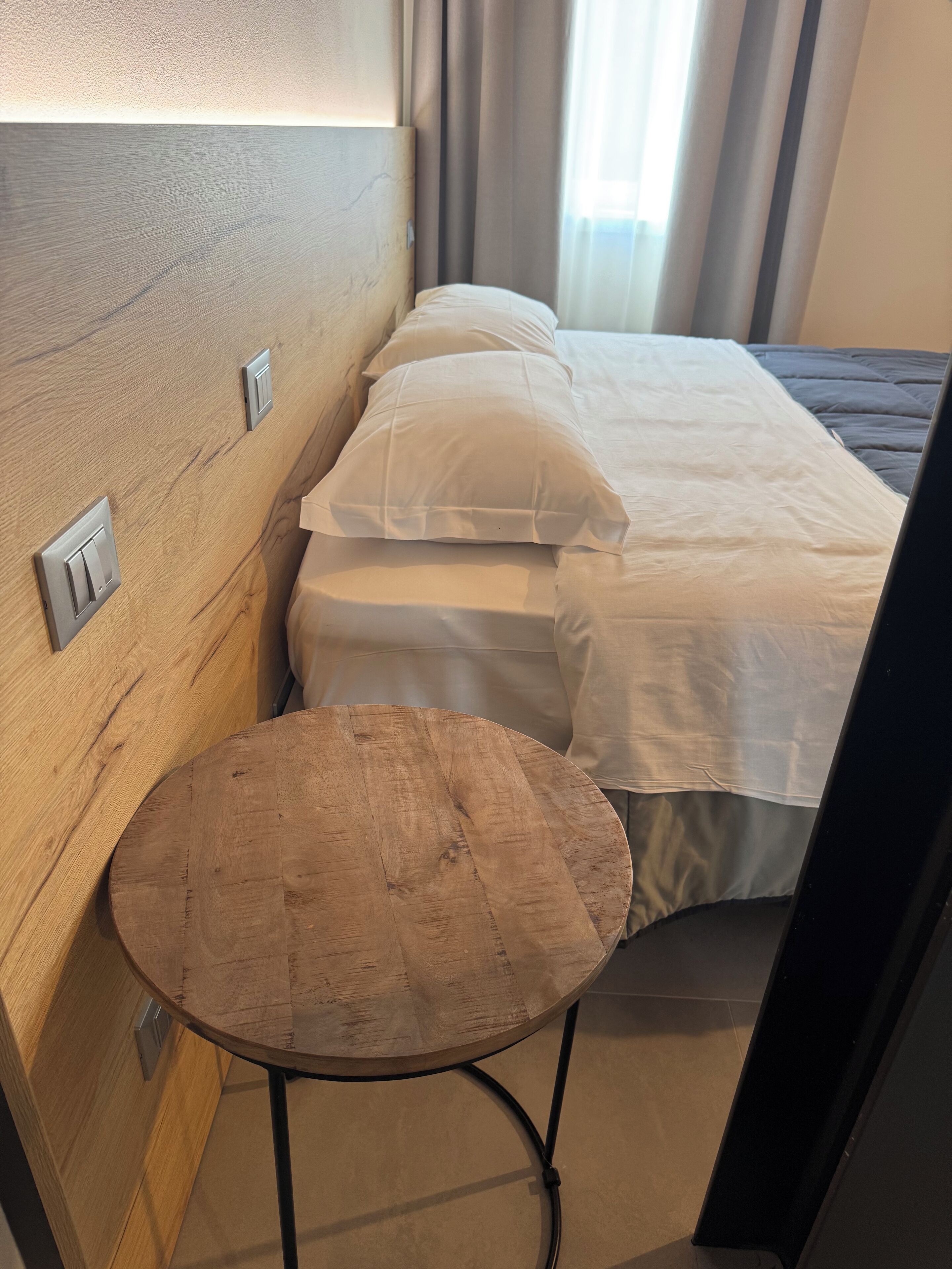 Makuuhuoneessa on puinen sängynpääty, pyöreä puinen pöytä, valkoisilla vuodevaatteilla varustettu sänky ja ikkuna, jossa on verhot.