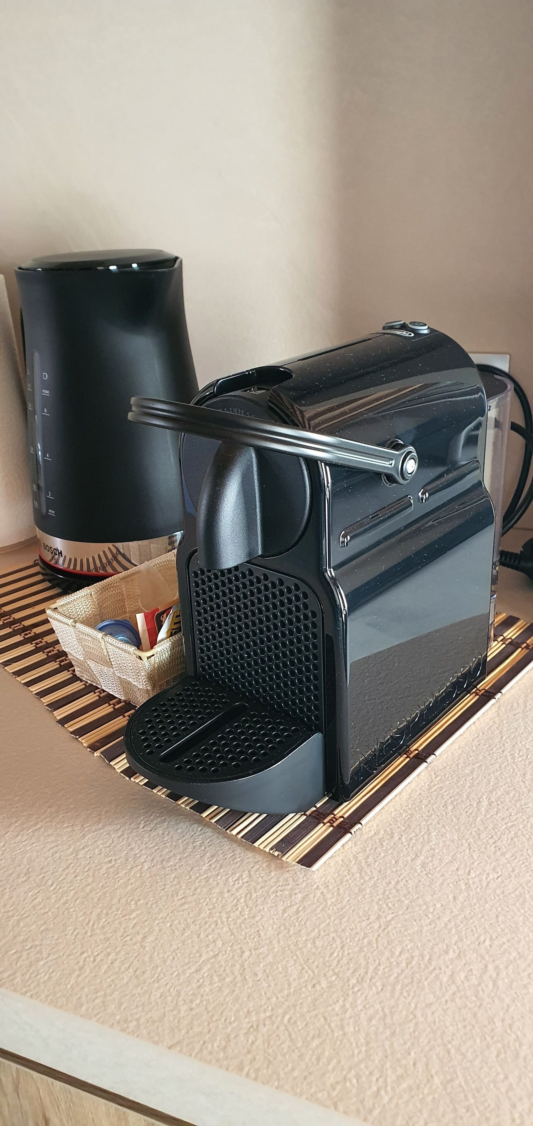 A black coffee maker on a bamboo mat with a woven basket containing books and a red object.