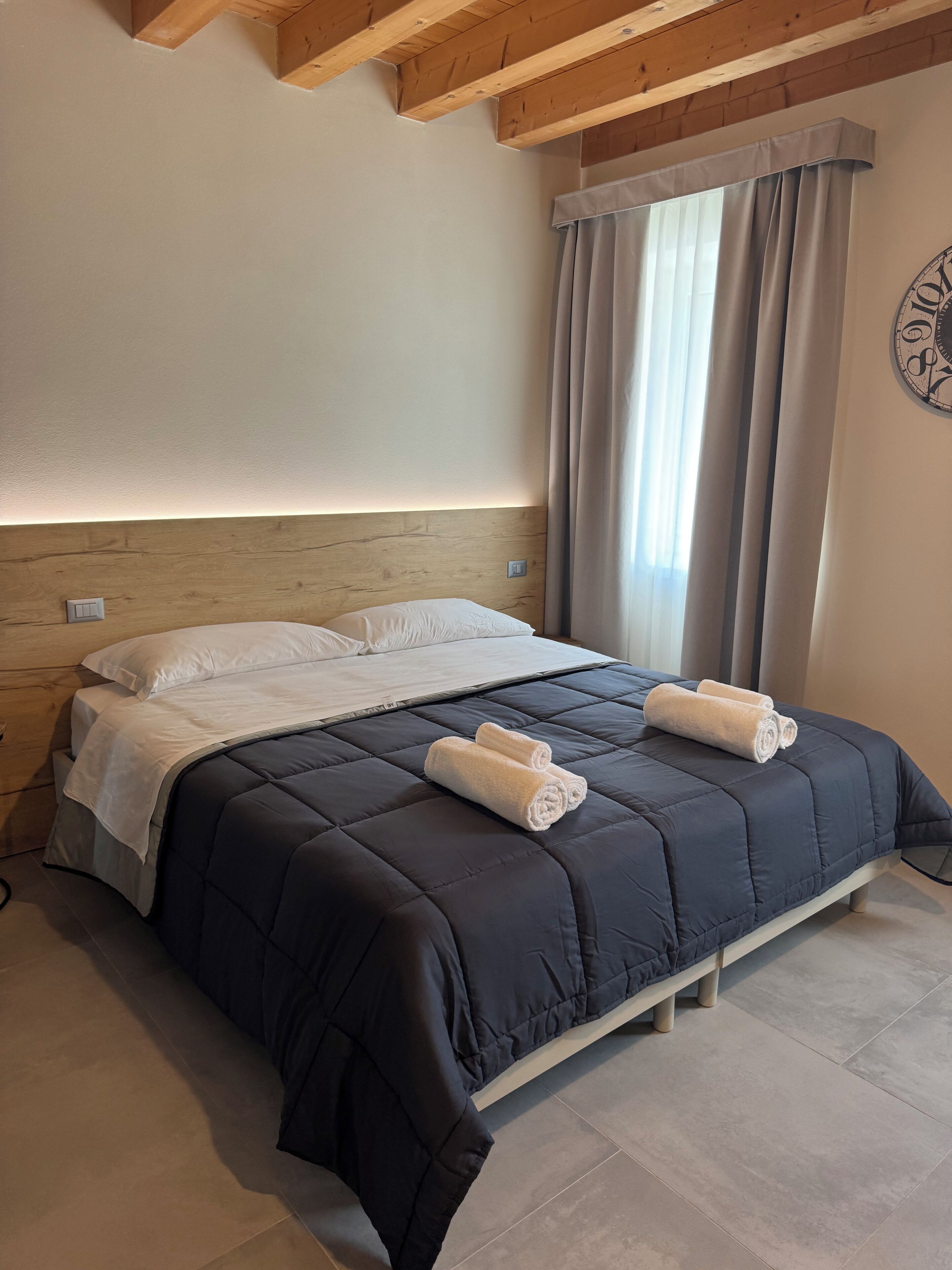 A bedroom with a dark blue bedspread, two rolled towels, wooden headboard, and a window with curtains.