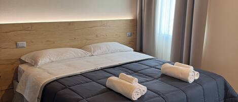 A bedroom with a dark blue bedspread, two rolled towels, wooden headboard, and a window with curtains.