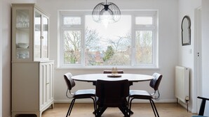 A dining area with a round table and four chairs, a white cabinet with glass doors, a large window with a view of trees and buildings, and a black pendant light. - GuestReady - Charming 3-Bedroom Apartment in NW11 (London)