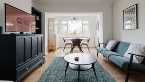 A modern living room with a TV, a sofa, a dining table, and a kitchen area. - GuestReady - Charming 3-Bedroom Apartment in NW11 (London)