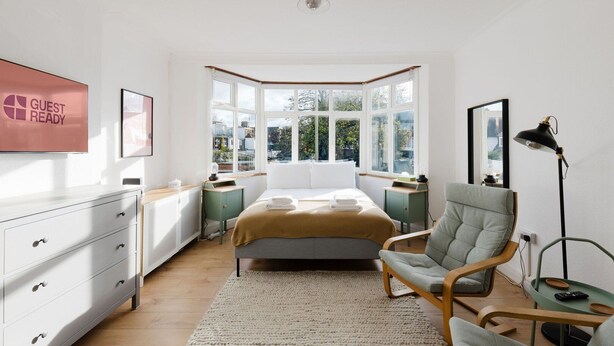 A bedroom with a bed, a chair, a desk, a lamp, and a window with a view of buildings. - GuestReady - Charming 3-Bedroom Apartment in NW11 (London)