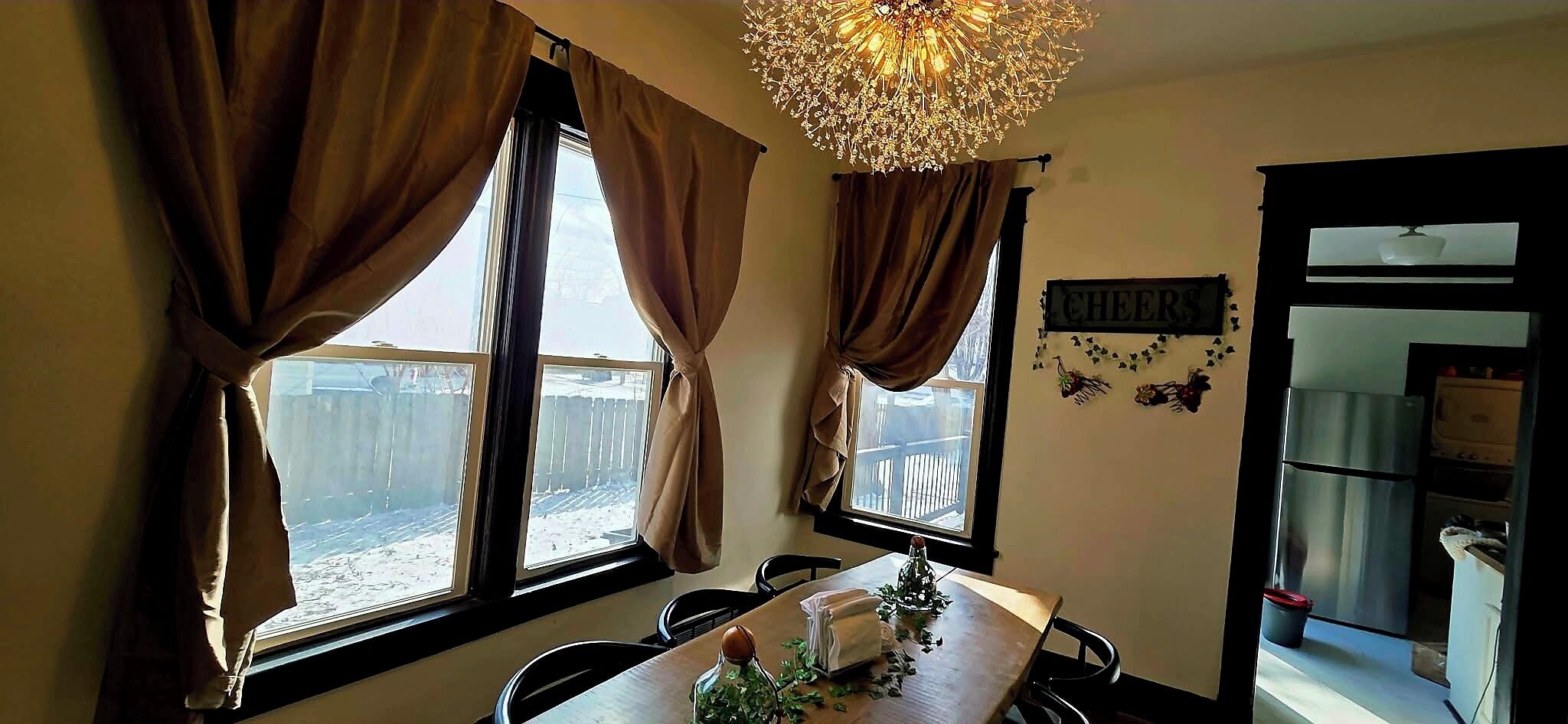 Un espace repas comprenant une table, des chaises, un lustre et une vue sur une terrasse en bois, visible à travers de grandes fenêtres.