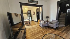 Un ampio soggiorno con parquet, due poltrone, un tavolino da caffè e una scala. Un cartello con la scritta 'WELCOME' è appeso sopra la porta che conduce alla sala da pranzo.