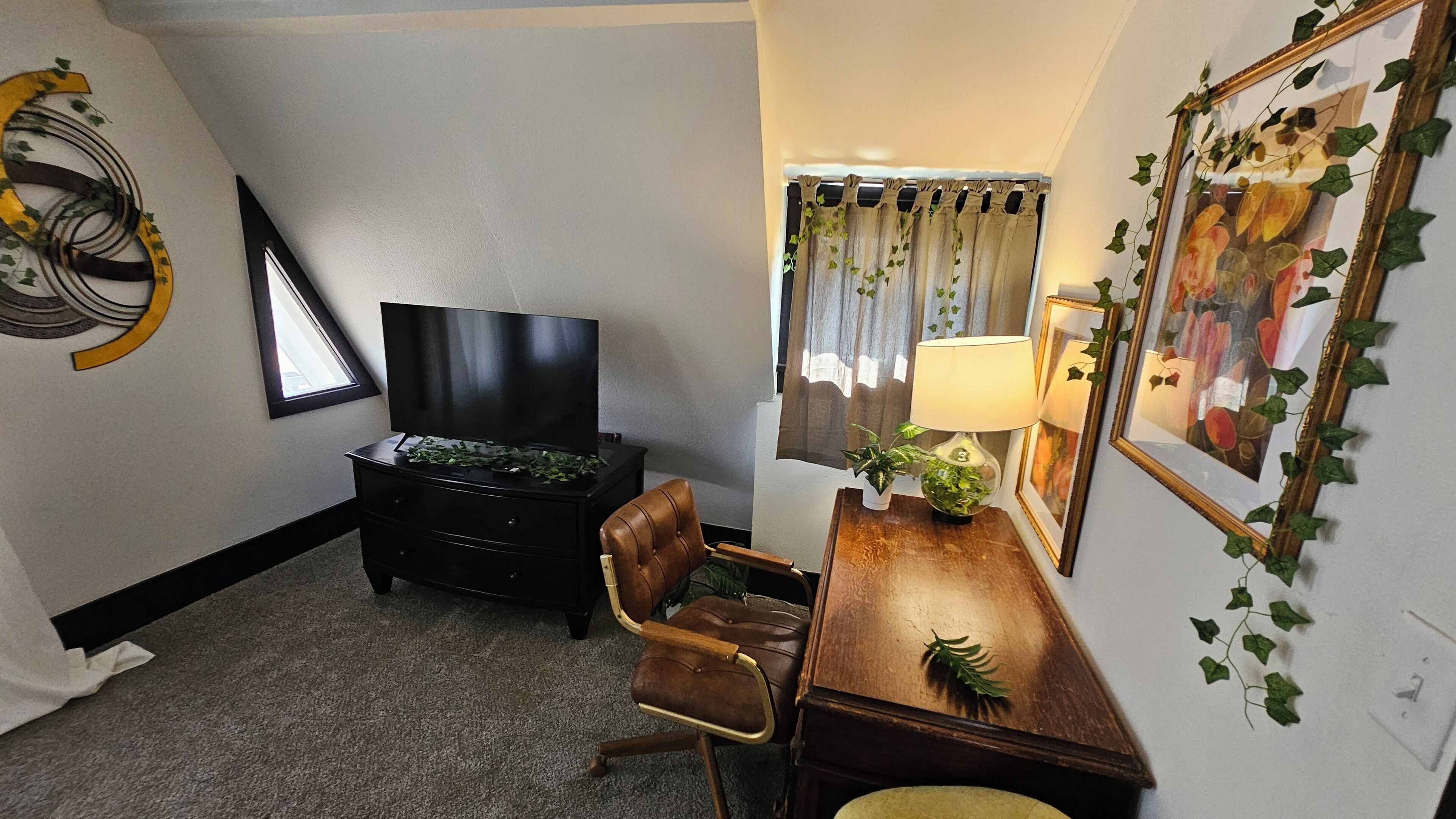 Een woonkamer met een flatscreen televisie, een bruine leren fauteuil, een houten bijzettafeltje met een lamp, en een raam met gordijnen.