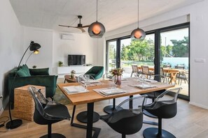 Ein moderner Essbereich mit einem Holztisch, gedeckt für vier Personen, umgeben von schwarzen Stühlen. Ein Fernseher ist an der Wand montiert, und das Zimmer verfügt über große Fenster mit Blick ins Freie.