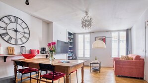 Uma sala de estar moderna com uma mesa de jantar de madeira, cadeiras, um relógio de parede grande, uma estante, um sofá e um lustre.