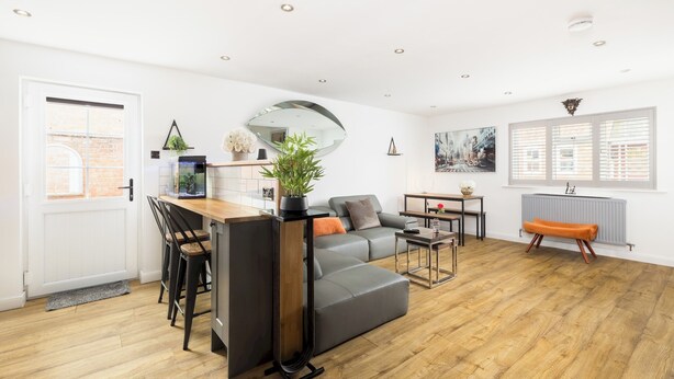 A modern living room with a grey sofa, wooden flooring, and a dining area with a black chair and table. - Stylish & Modern 2 Bed with Parking - Pass the Keys (Stratford-upon-Avon)
