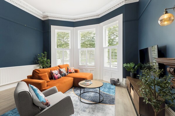 Una habitación con pared azul oscuro, ventana saliente con marco blanco, banco de madera y sofá gris con cojines de colores.