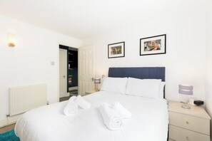 Una cama bien hecha con sábanas blancas y dos toallas enrolladas, una lámpara de noche y dos cuadros en la pared.