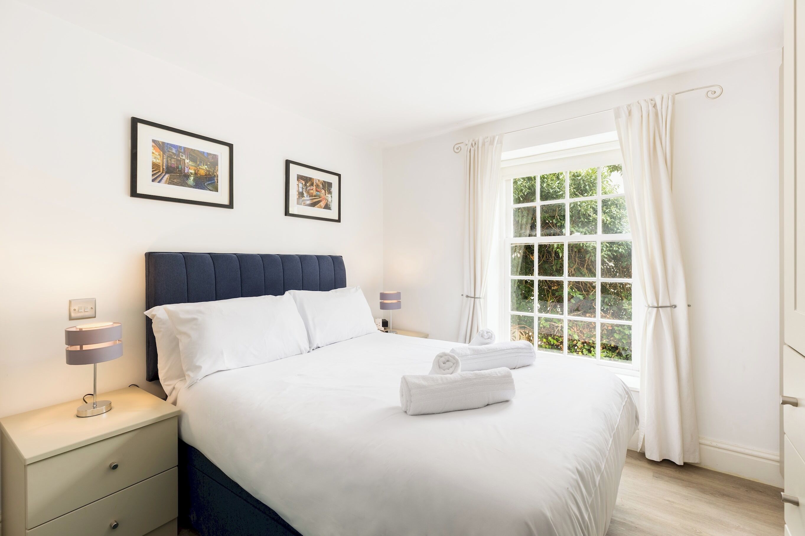 A neatly made bed with white linens and two rolled towels, a bedside lamp, and two framed pictures on the wall.