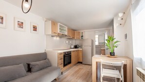 Een ruimtelijke leefruimte met een kitchenette, een bank, een eettafel met een stoel en een potplant.