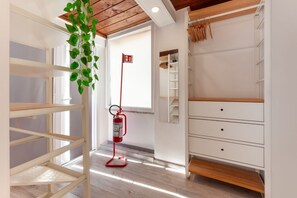 Ein Zimmer mit Holzbalkendecke, weißen Wänden, einem weißen Schminktisch und einem hängenden Topf mit Pflanze. Ein roter Feuerlöscher ist ebenfalls zu sehen.