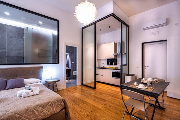 A modern hotel room with a bed, a dining table, a kitchen area, and a large mirror.