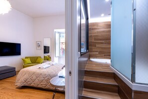 A modern bedroom with a bed, a grey sofa, a TV, and a bathroom with a bathtub and wooden flooring.