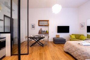 A modern bedroom with a bed, a desk, a chair, a television, and a small kitchenette.