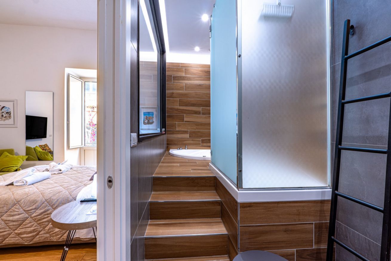 A modern hotel room with a bed, a small table, a shower, and a staircase leading to a balcony.
