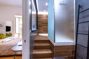 A modern hotel room with a bed, a small table, a shower, and a staircase leading to a balcony.