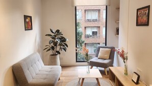 Een moderne woonkamer met een grijze bank, een klein bijzettafeltje, een potplant en uitzicht op een bakstenen gebouw door een groot raam.