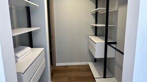 Een smalle inloopkast met witte schapkasters en lades, houten vloeren en een ingebouwd plafondlampje.