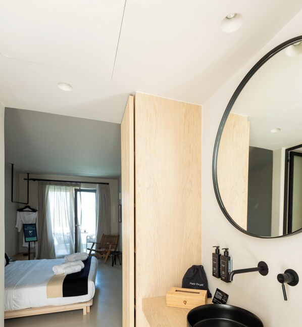 A modern hotel room with a bed, a wooden wardrobe, a round mirror, a sink, and toiletries. - Hotiday Mykonos South Coast (Mykonos)