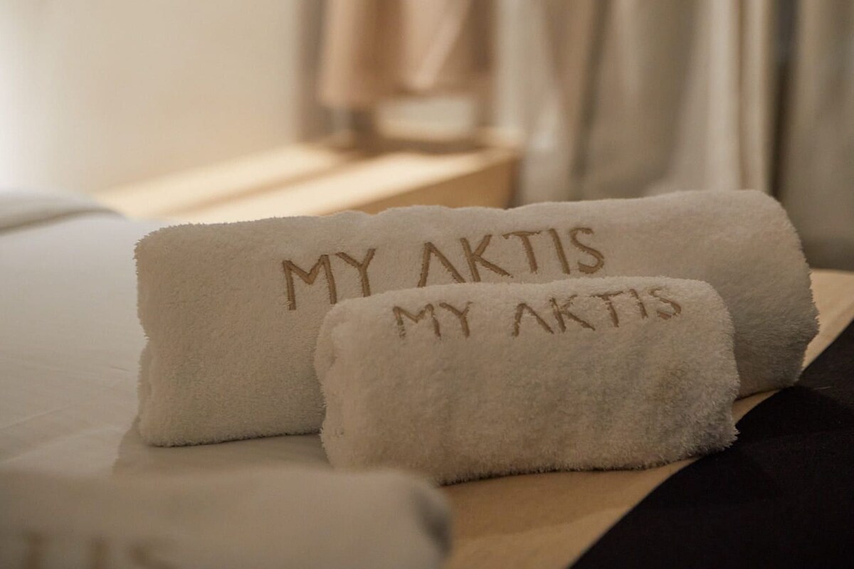 Une chambre d’hôtel avec un lit, deux serviettes brodées avec le logo « MY AKTIS », un peignoir suspendu à un porte-manteau et une fenêtre avec des rideaux.