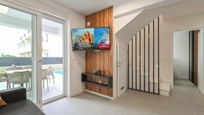 Ett modernt vardagsrum med en soffa, en tv som är monterad på en träpanel och utsikt över en pool utanför.