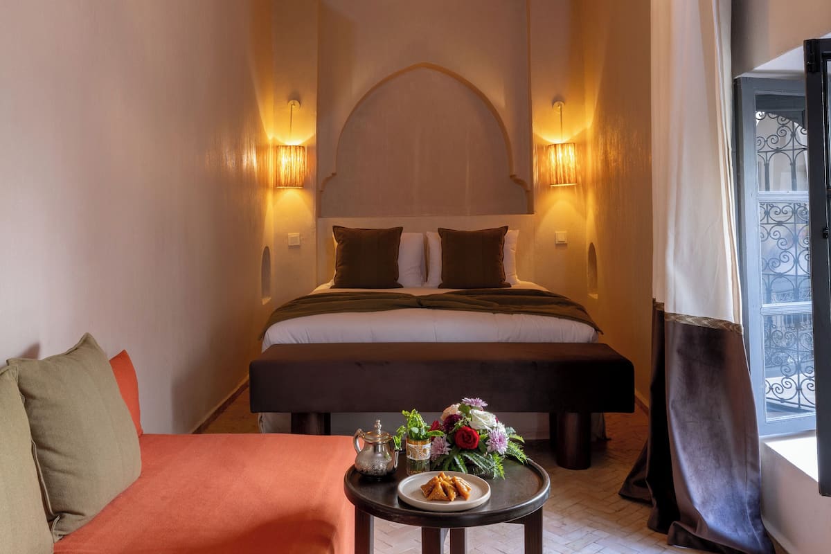 Una habitación de hotel con cama, sofá, una mesita con un arreglo floral y una ventana con cortinas.