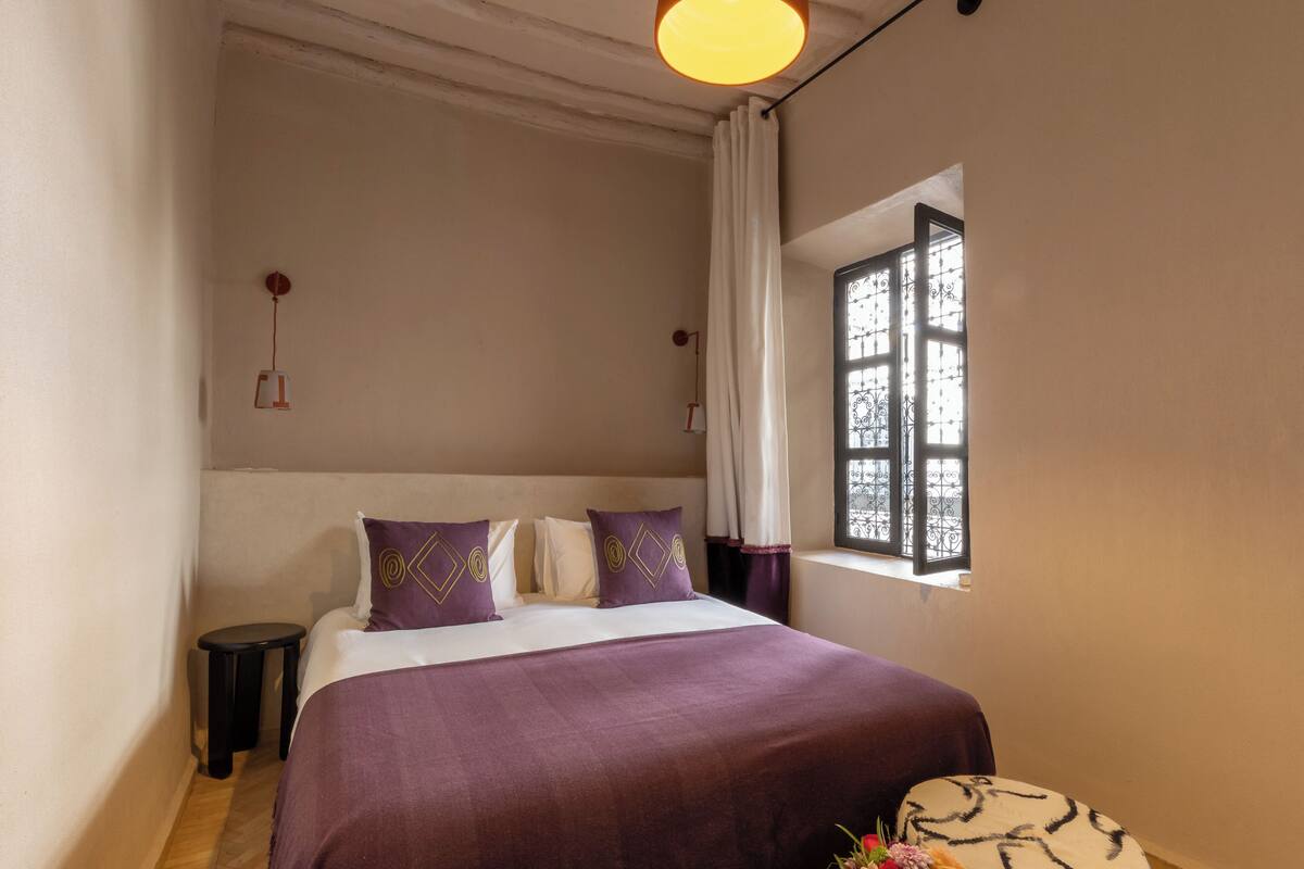Un dormitorio con cama, ventana con vistas, una mesita y una lámpara de pared.