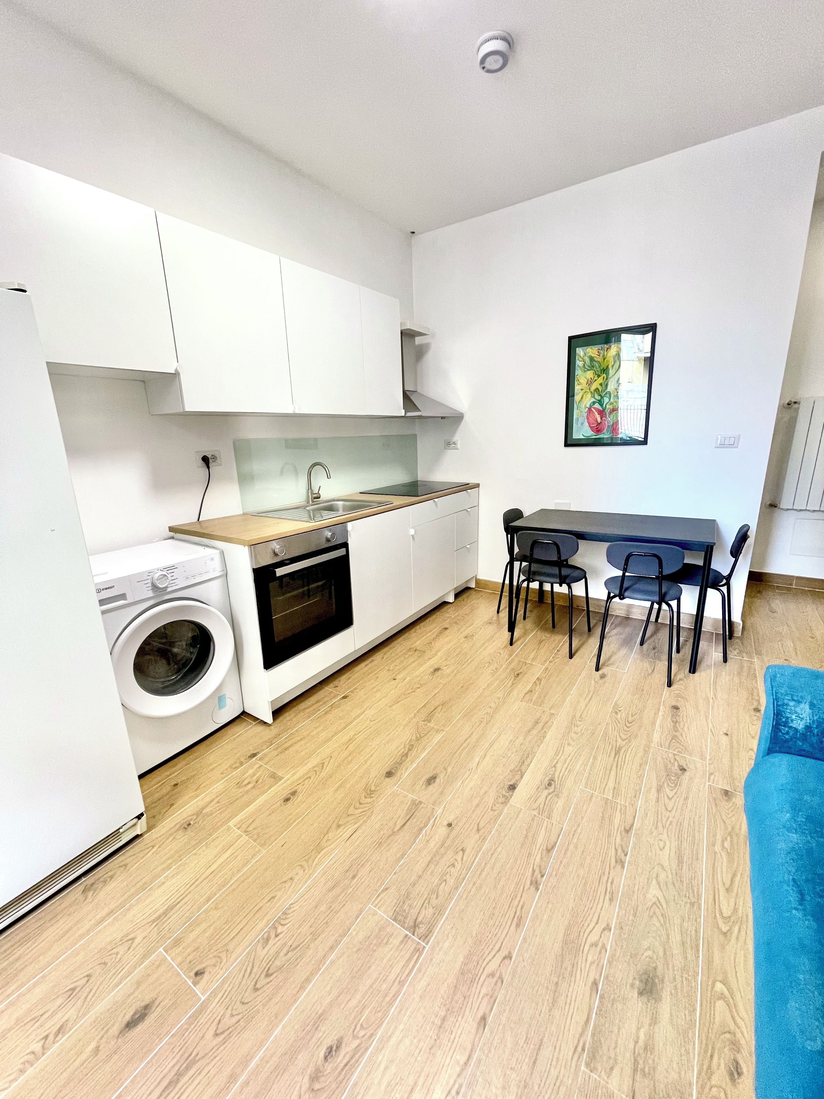 Eine kompakte Küche mit Waschmaschine, ein Essbereich mit Tisch und Stühlen sowie ein blaues Sofa.