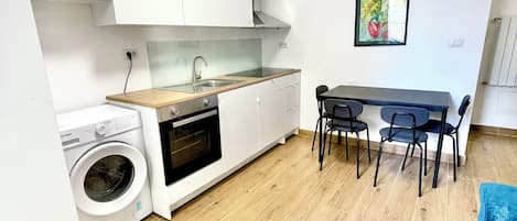A compact kitchen with a washing machine, a dining area with a table and chairs, and a blue sofa.
