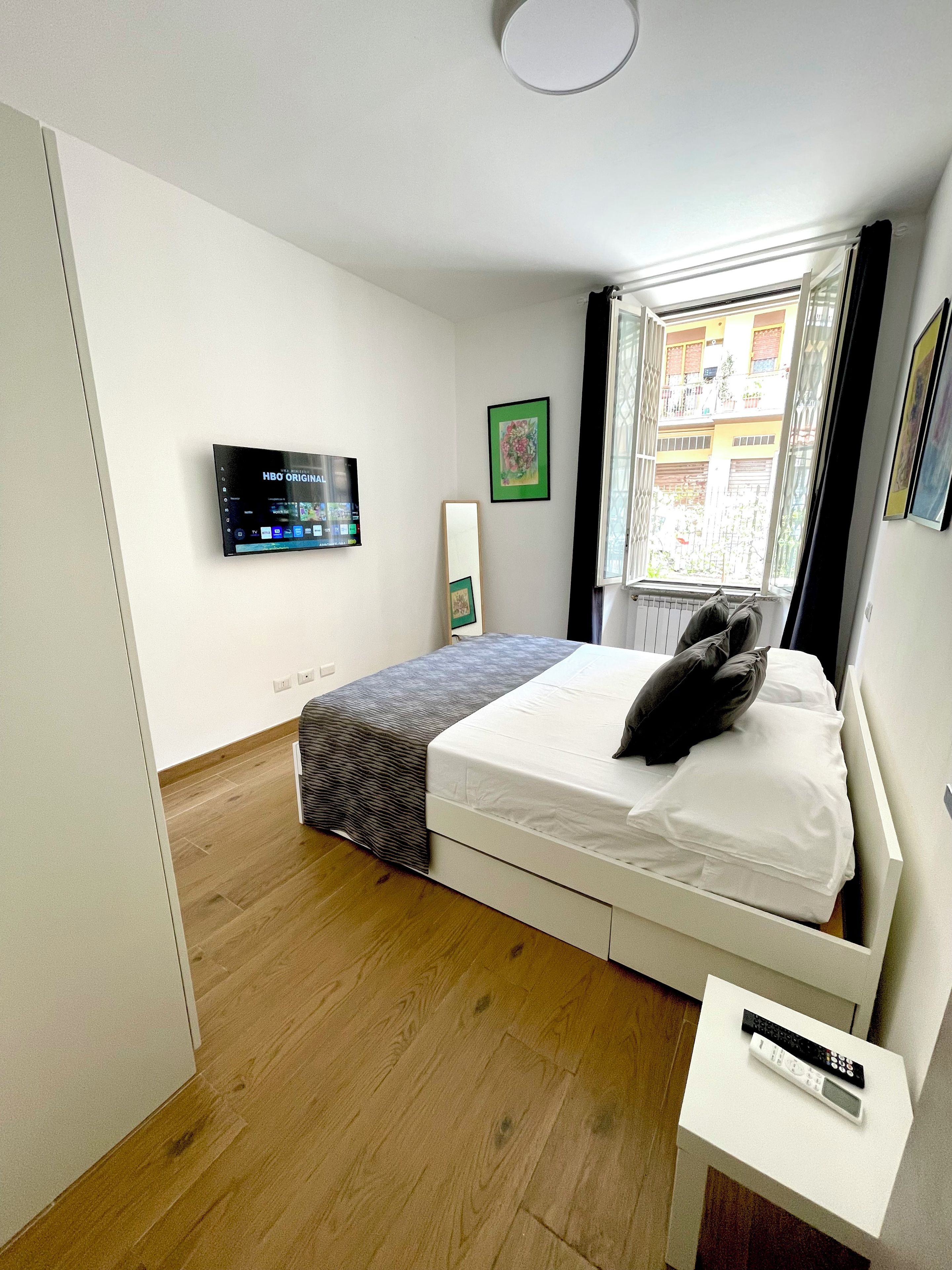 A bedroom with a bed, a TV mounted on the wall, a window with curtains, and a bedside table with a remote control.