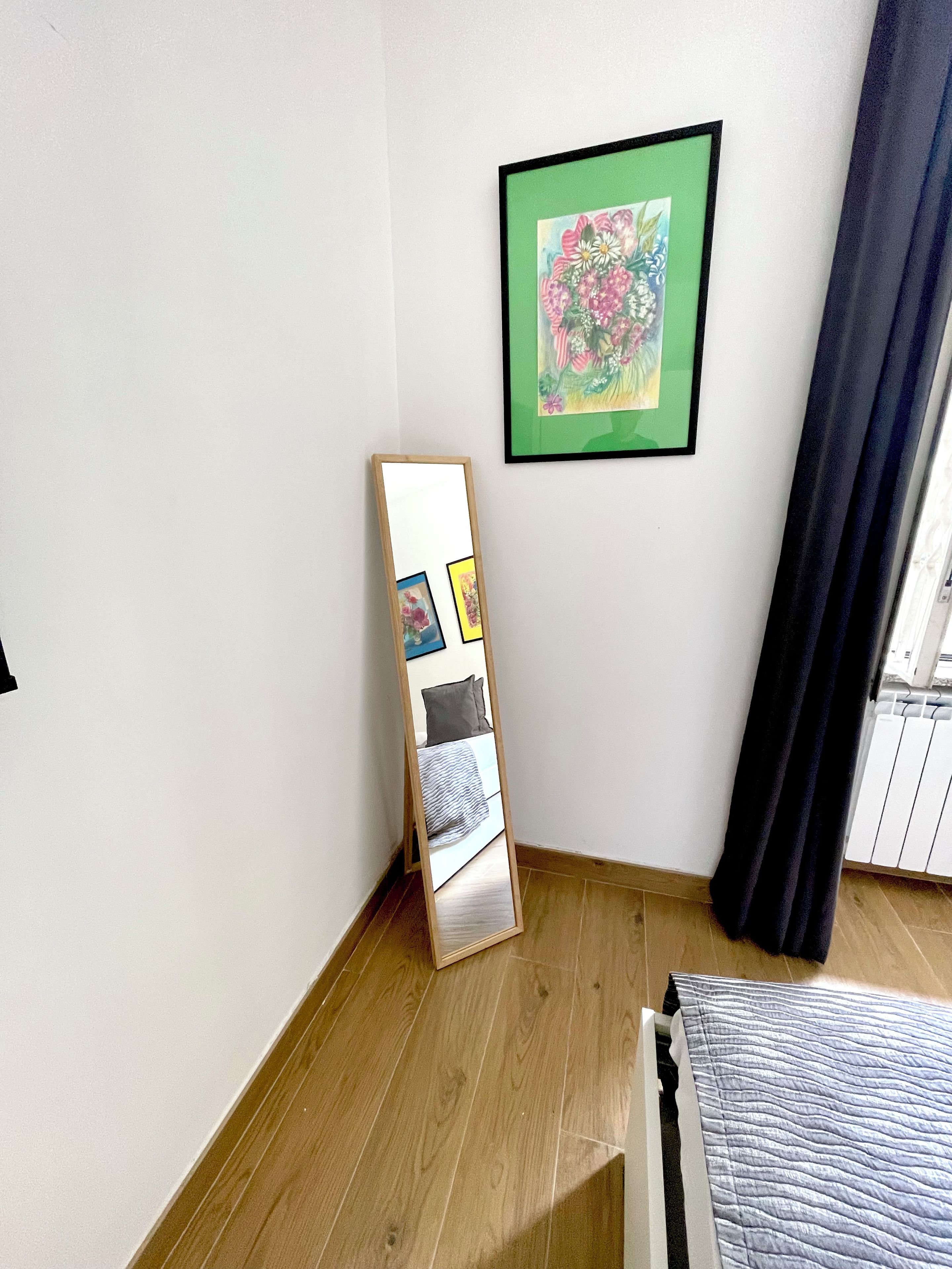Ein Zimmer mit einem Bett, einem an der Wand lehnten Spiegel, einem gerahmten Blumenbild und einem Vorhang vor dem Fenster.