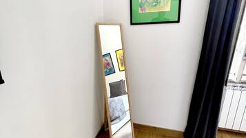 Ein Zimmer mit einem Bett, einem an der Wand lehnten Spiegel, einem gerahmten Blumenbild und einem Vorhang vor dem Fenster.