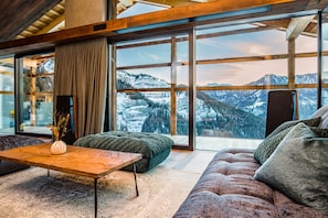 Un soggiorno moderno con un'ampia finestra che offre vista sulle montagne, un tavolino in legno, un divano verde e un vaso decorativo.