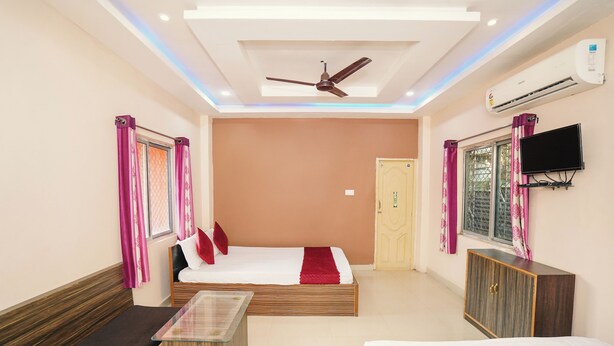 A hotel room with a bed, a sofa, a small table, a ceiling fan, an air conditioner, and a TV mounted on the wall. - PURNI HOTEL  (Kanthi)