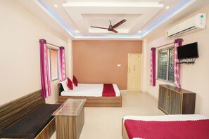 A hotel room with a bed, a sofa, a small table, a ceiling fan, an air conditioner, and a TV mounted on the wall. - PURNI HOTEL  (Kanthi)