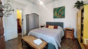 A bedroom with a bed, a wooden table, a wardrobe, a painting, and a chandelier.