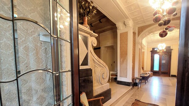 A grand hallway with a marble floor, a wooden chair, a small table, and a staircase with a decorative vase.