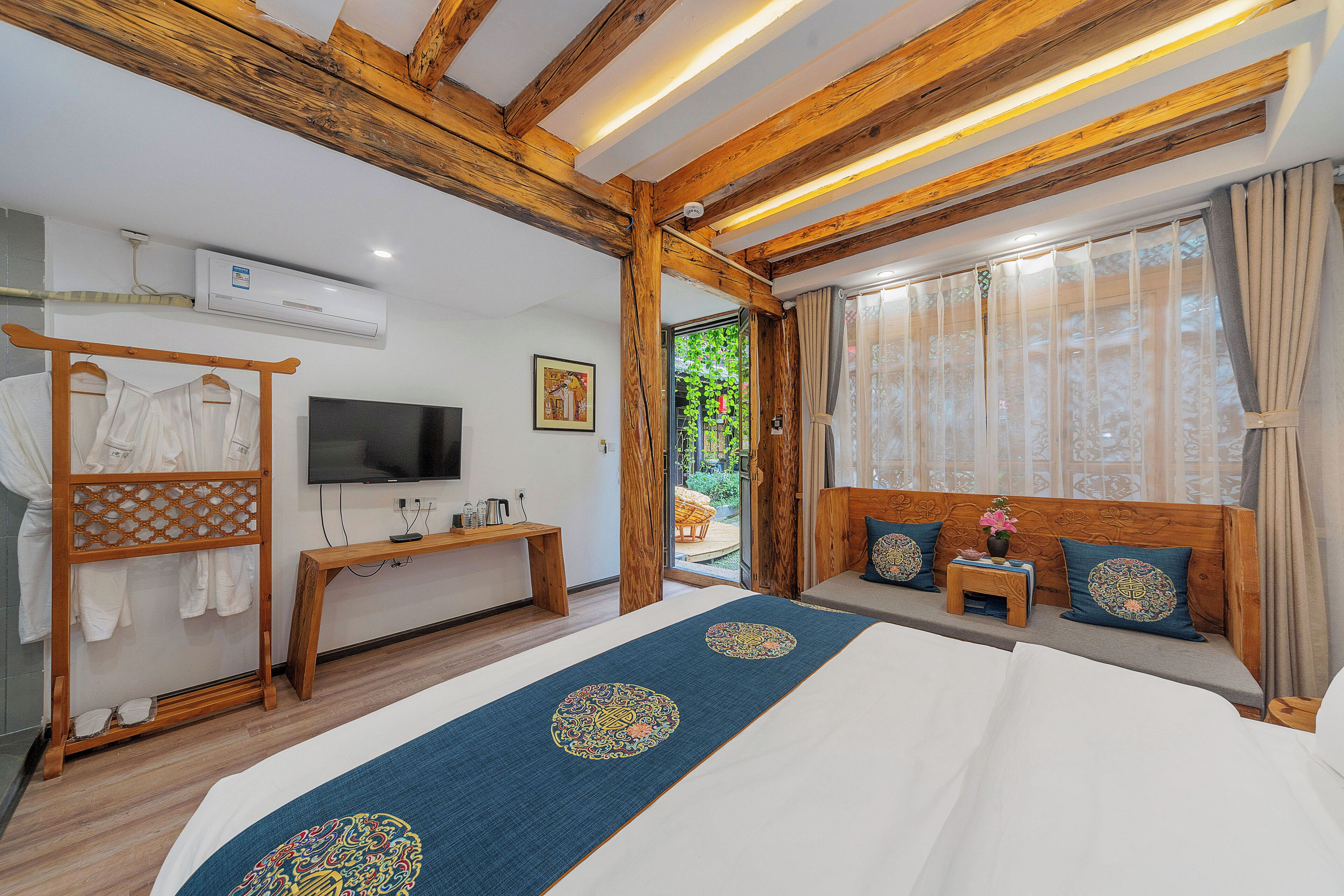 A hotel room with a wooden ceiling, a bed with a blue runner, a TV, a bench, and a window with curtains.