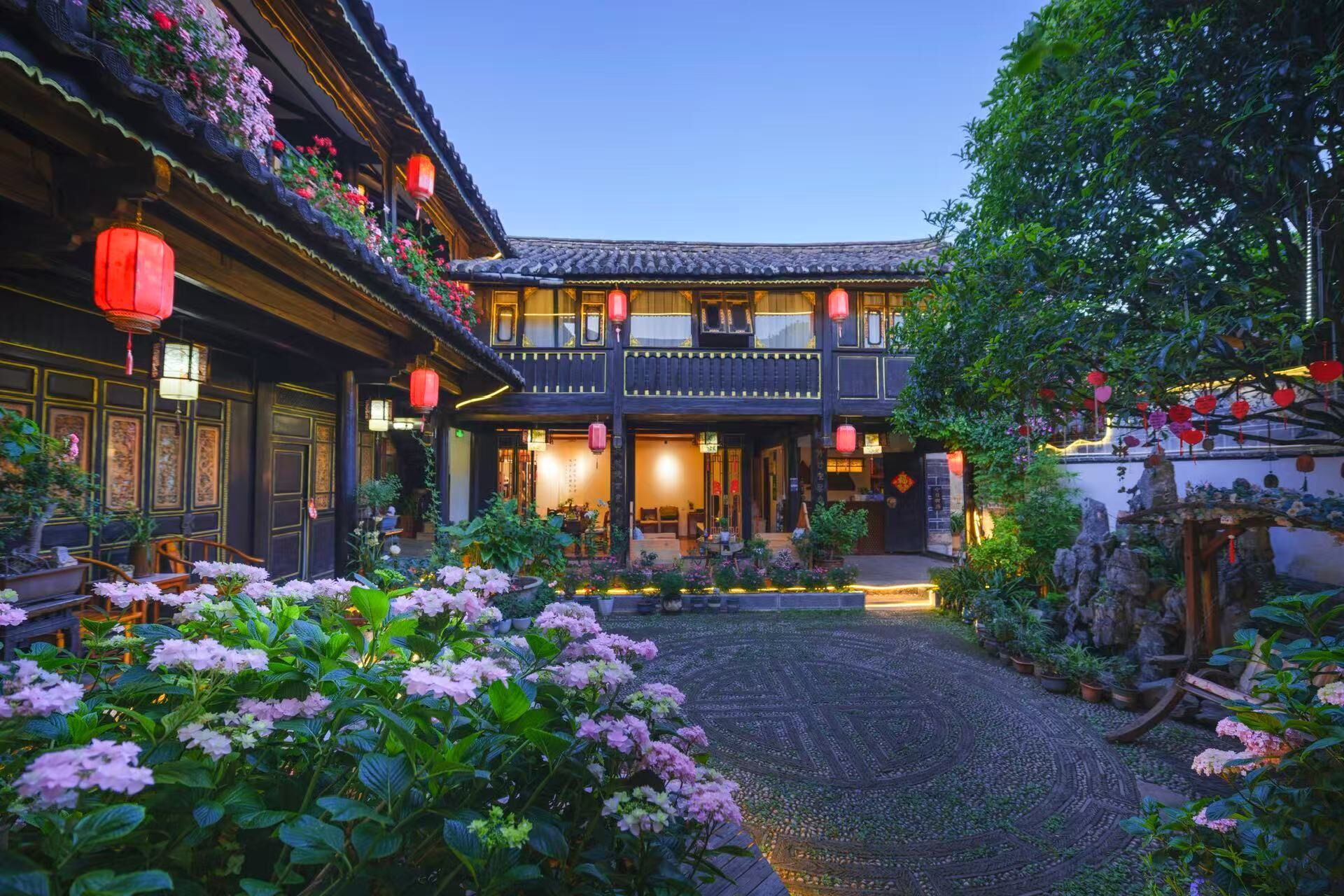 A traditional courtyard with a wooden building, lanterns, and flowering plants.
