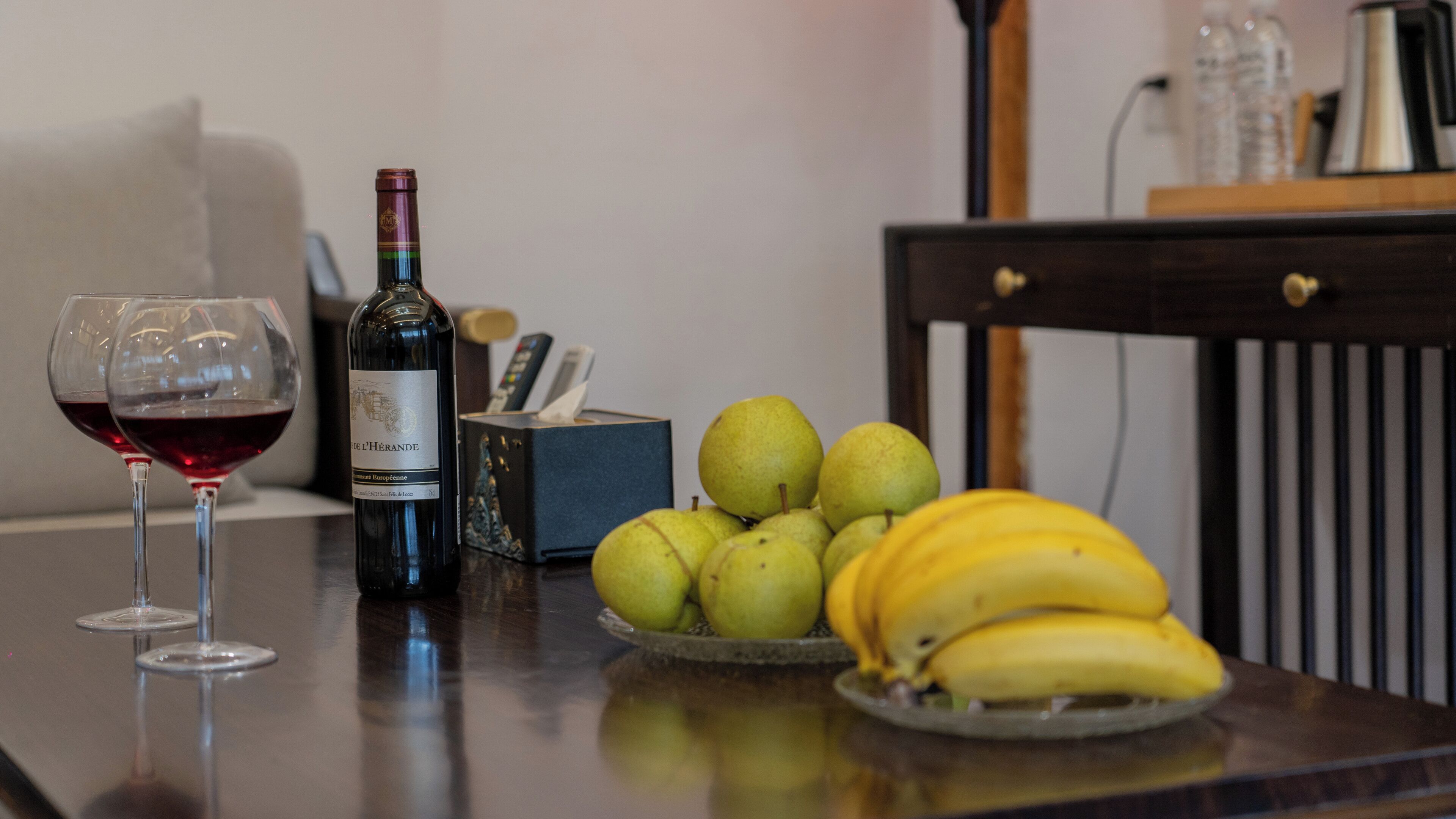 Ein Tisch mit zwei Weingläsern, einer Weinflasche, einer Taschentuchbox und einer Schale mit Bananen und Birnen.