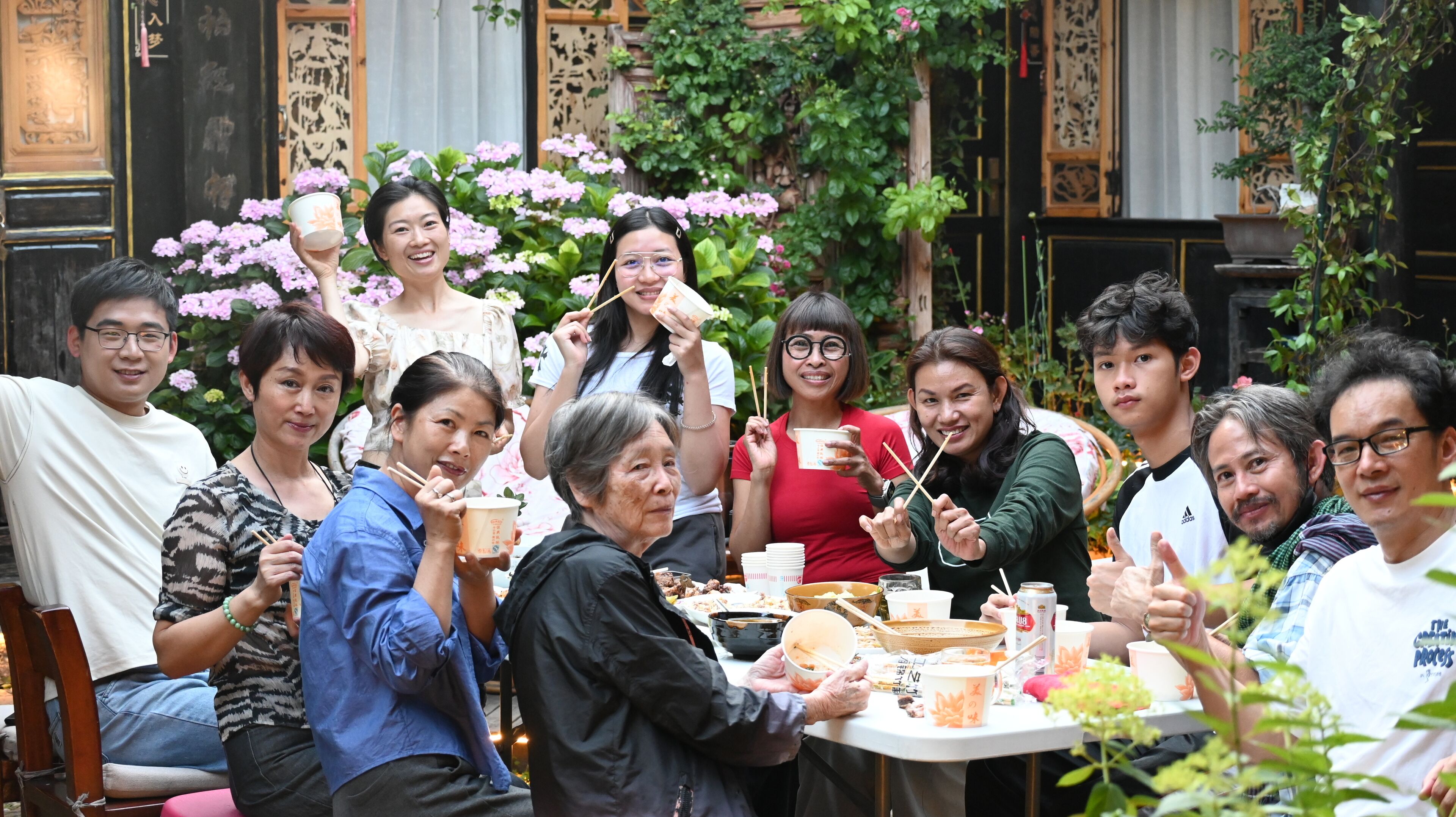 A group of people seated around a table with food and drinks, in an indoor setting with traditional decor and plants.