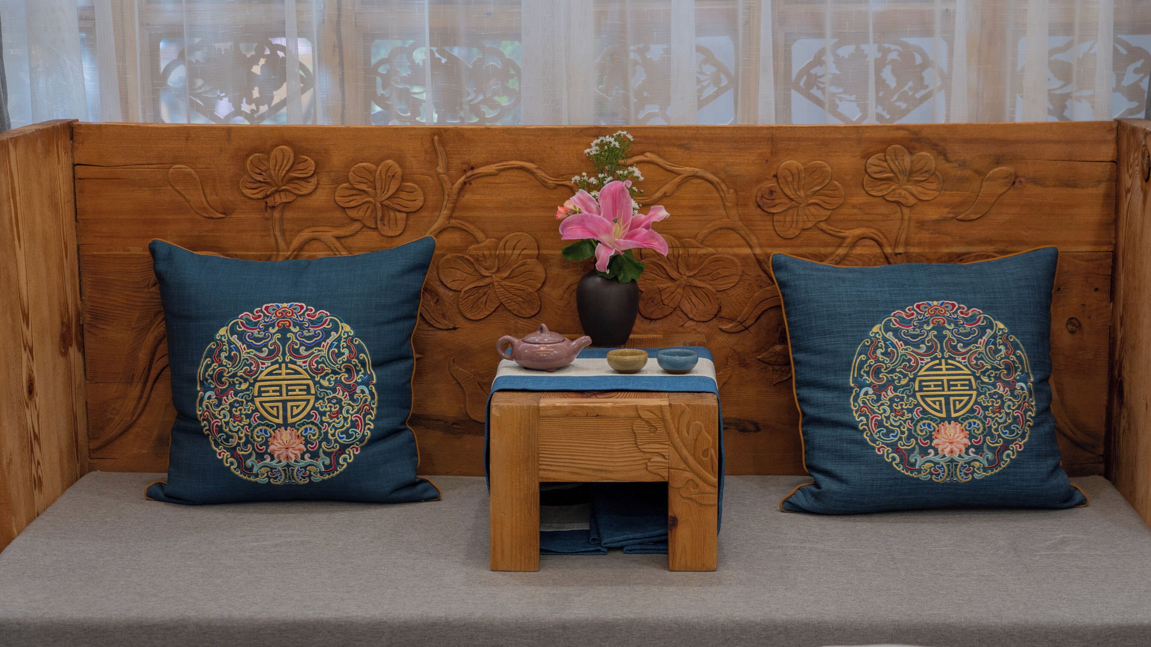 A wooden sofa with intricate carvings, two decorative pillows with embroidered designs, a small wooden table with a vase of flowers, and a teapot.