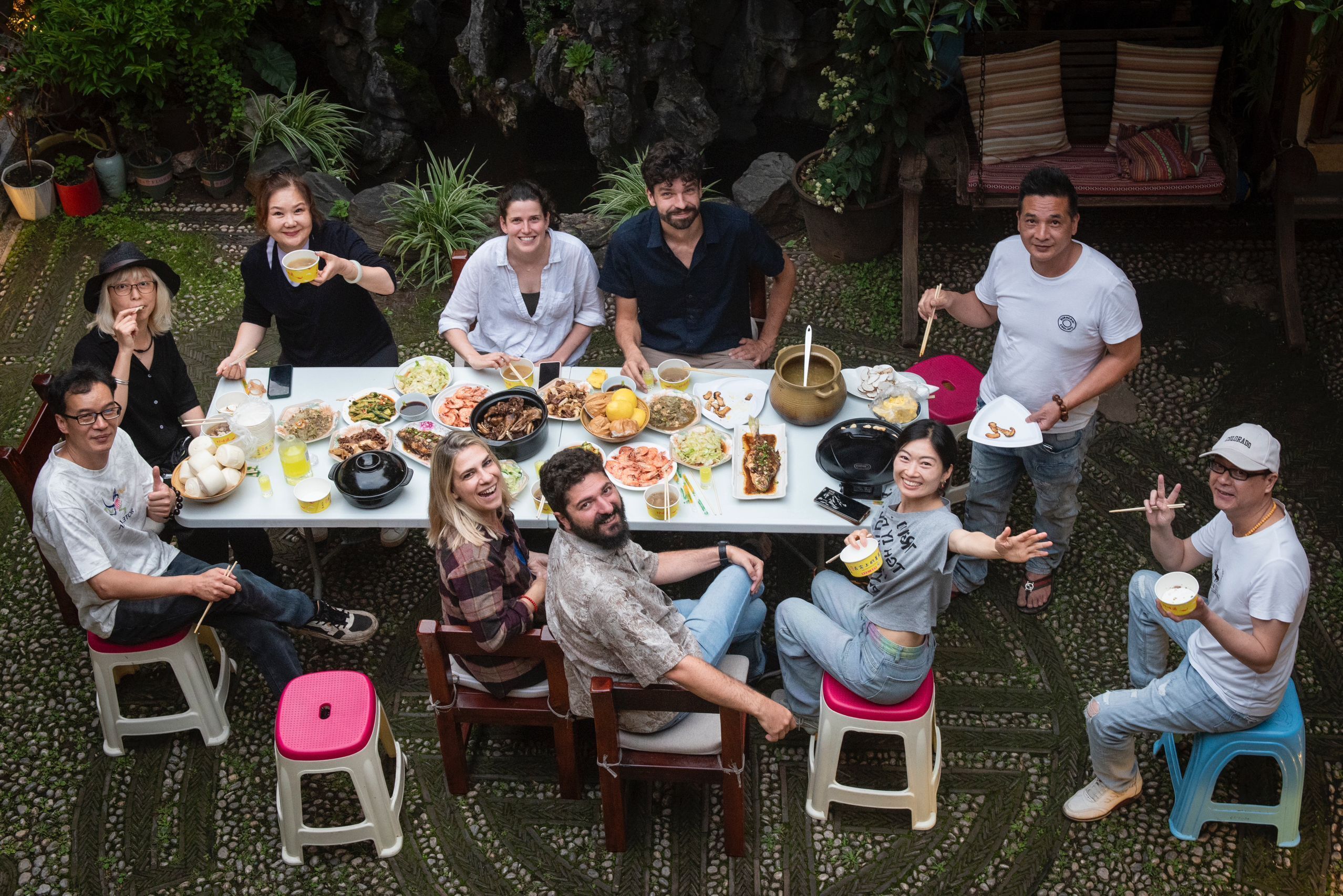 A group of people seated around a table with various dishes, in an outdoor setting with plants and a rock formation in the background.