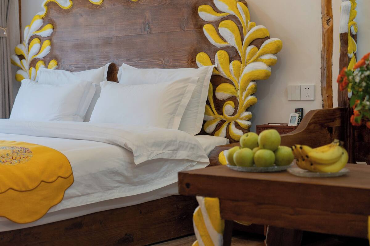 Un lit bien fait, avec du linge de lit blanc et un coussin décoratif jaune. Une tête de lit en bois ornée de motifs de feuilles jaunes et blanches est visible. Une table de chevet en bois, sur laquelle se trouve une assiette de bananes et de pommes, est placée devant le lit.