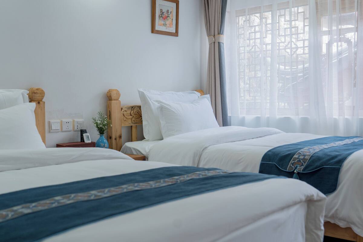 Une chambre d’hôtel avec deux lits, une tête de lit en bois, une table de chevet avec un vase et une fenêtre ornée de rideaux légers.