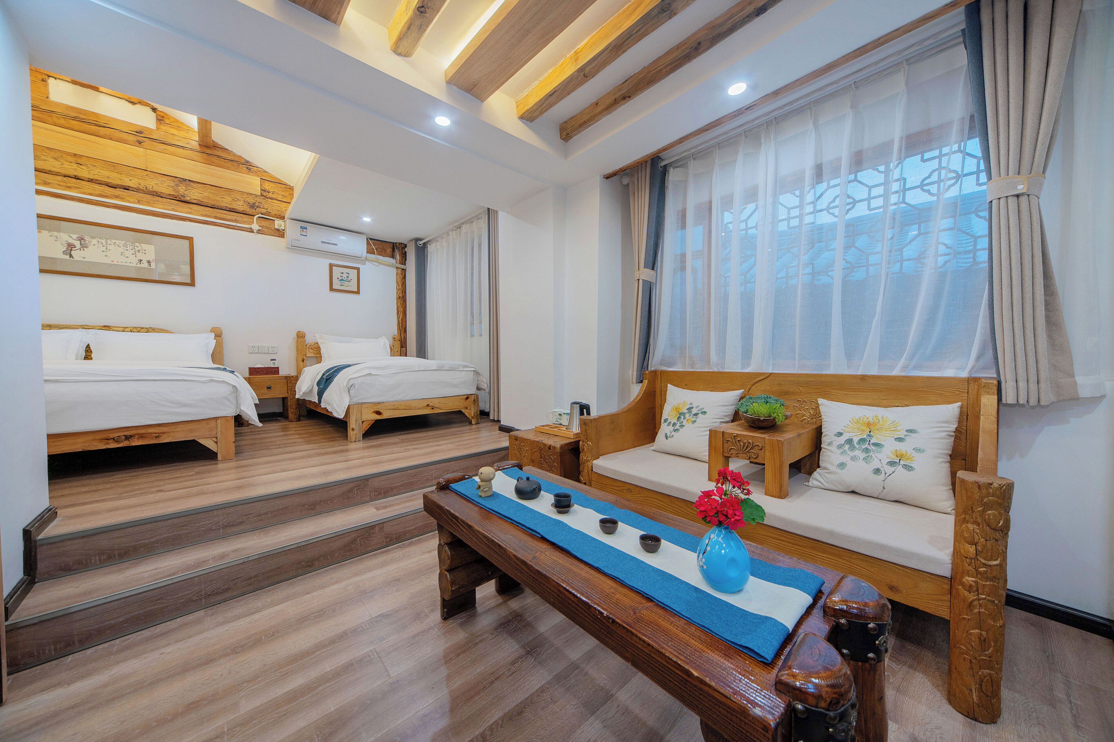 Habitación de hotel con dos camas de madera, un sofá, una mesa de centro con un jarrón y una ventana con cortinas traslúcidas.