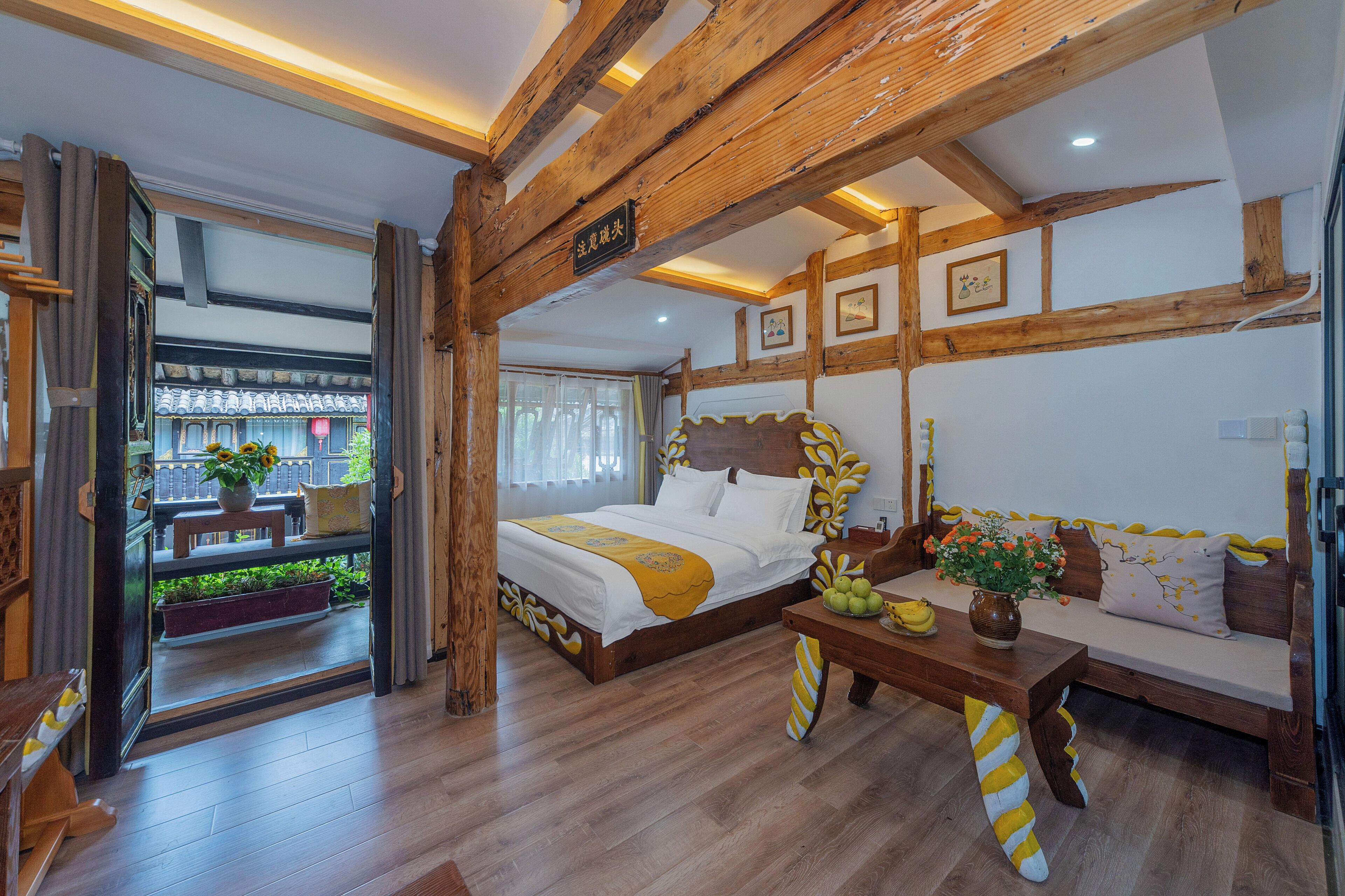 A room with a wooden ceiling, a bed with a yellow and white bedspread, a wooden bench, a table with a vase of flowers, and a view of a balcony with plants.